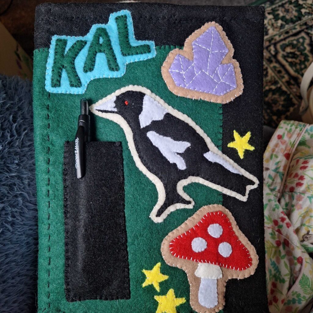 A green-and-black journal cover with handmade patches stitched on. There's a patch of a purple cluster of crystals, an Australian magpie and a red mushroom, plus three little yellow stars and a pen pocket. The top left patch reads KAL.