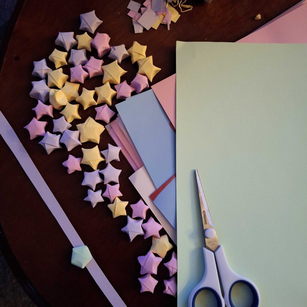 A group of colourful tiny paper stars lays against a wooden coffee table as seen from above. Next to the paper stars lays a stack of colourful pastel paper and a pair of scissors.