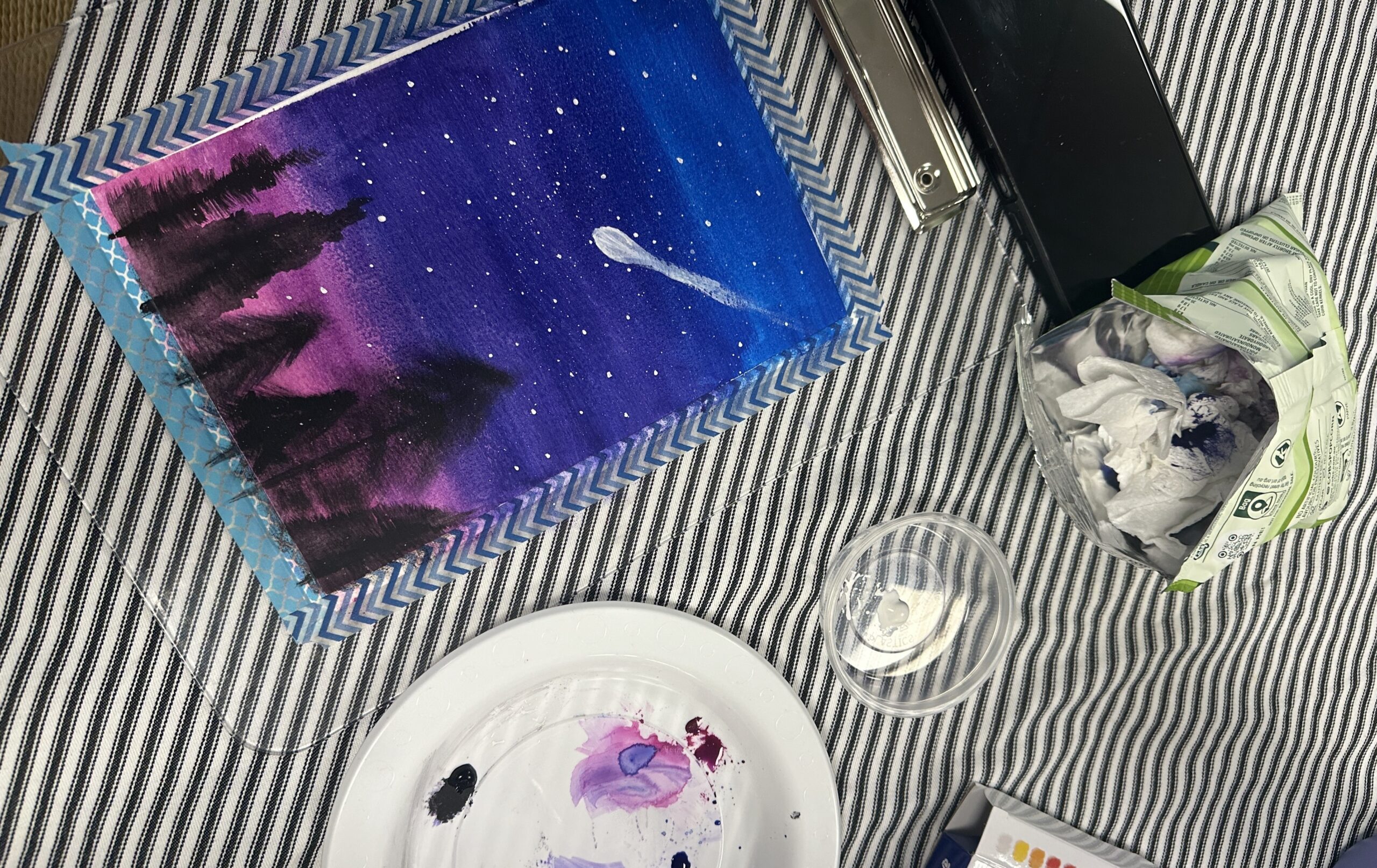 A painting of the night sky with a shooting star going through it. The sky is made up of shades of blue, purple and pink, and the silhouettes of pine trees line the bottom. The painting is laid against a table on a stripy tablecloth.