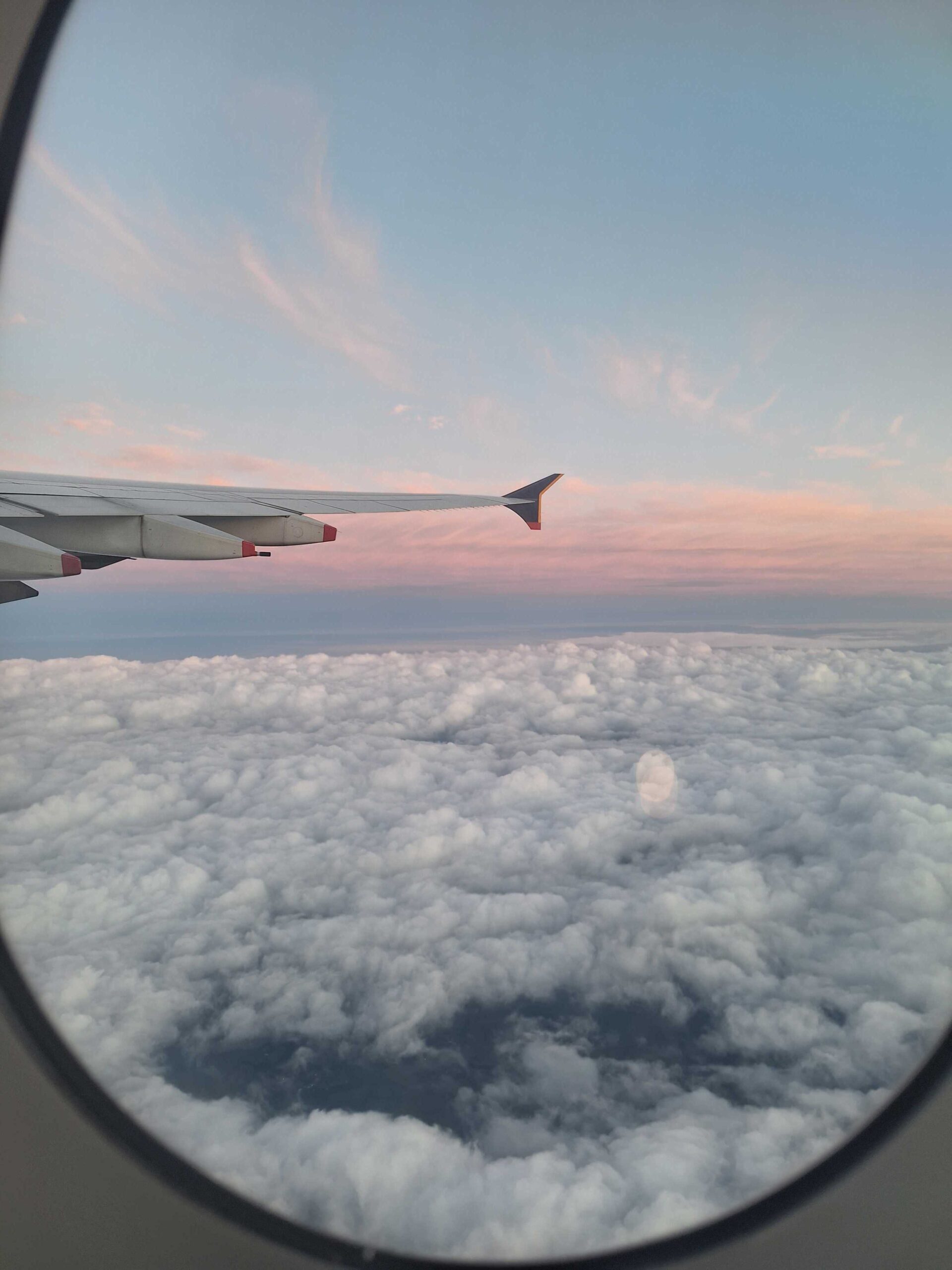 The view from a plane window. There's a soft pink/blue gradient in the sky that suggests the sun is setting. The window of the plane sticks out from the left. The plane is flying above a landscape of fluffy clouds.