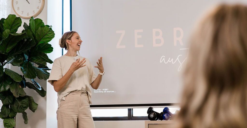 A woman stands in front of a projector with the Zebras Australia logo on it. She looks towards the projector and gestures with her hands in a way that implies she's speaking to a room full of people. Beside the projector stands a tall, leafy potted plant.