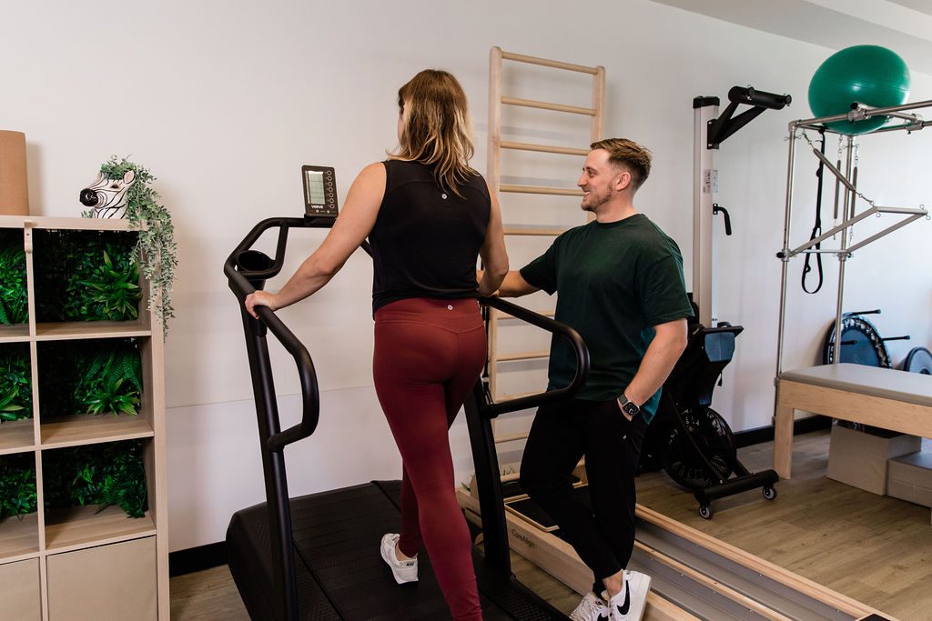 A man standing beside a woman on a treadmill in a light-filled gym space.