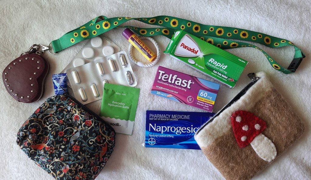 A spread of various small pharmaceutical and helpful items laid against a white blanket. The spread includes two small pouches, a sunflower lanyard, various over-the-counter medications, electrolytes and lip balm.