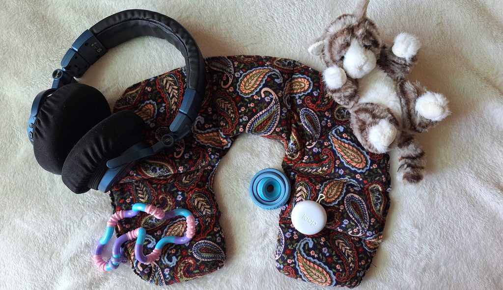 Various comfort items laid against a white blanket. They include a pair of blue headphones, a wheat bag shaped like a neck pillow, a small plushie of a cat, two fidgets and a white case for earplugs.