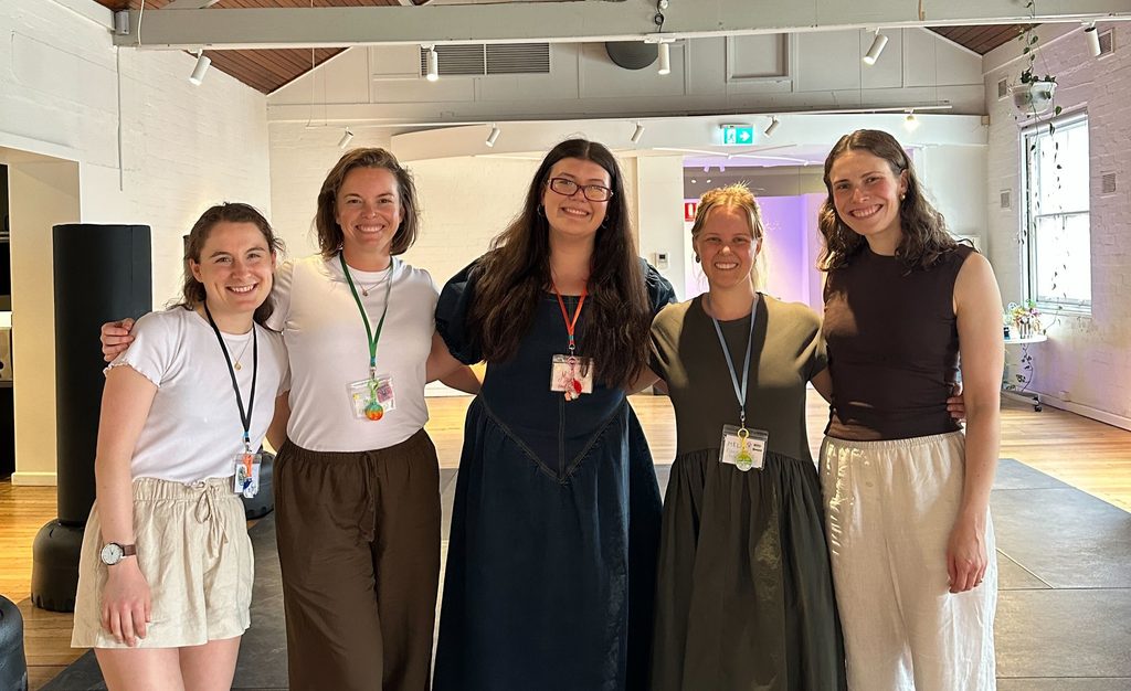 Sophia, Melanie, Manon, Mel and Lee from the Zebras team pose for a photo, smiling and with their arms around each other. They stand in a large hall where Conscious Combat Club activities take place.