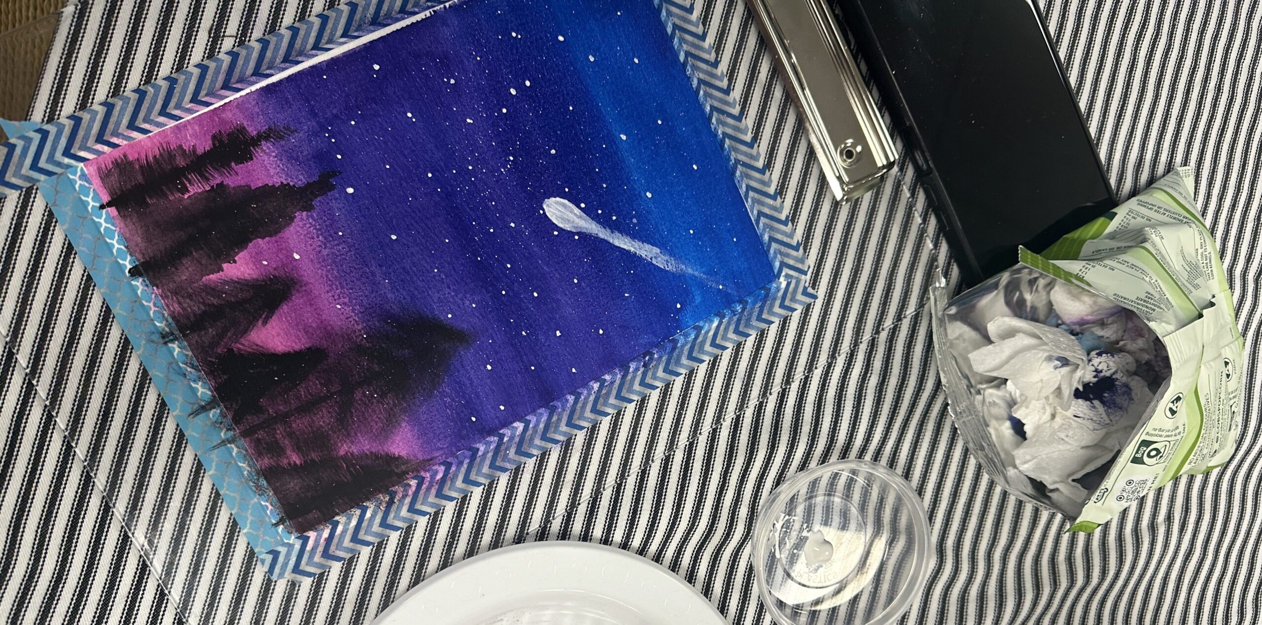 A painting of the night sky with a shooting star going through it. The sky is made up of shades of blue, purple and pink, and the silhouettes of pine trees line the bottom. The painting is laid against a table on a stripy tablecloth.