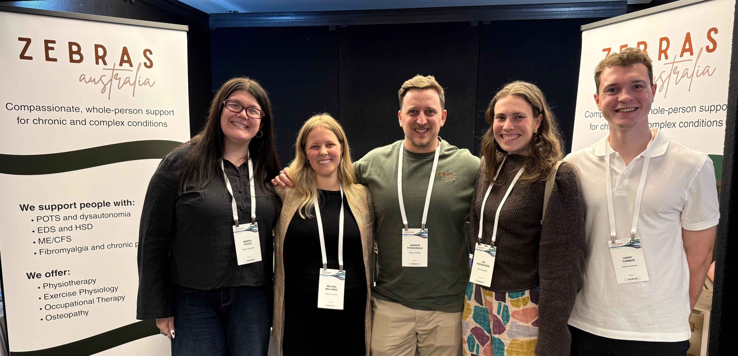 Manon, Mel, Andrew, Lee and Harry pose for a photo in front of the Zebras Australia stand at the Australian POTS Conference in 2026. They Stand beside a large standing banner that reads Zebras Australia, along listing the conditions we support and the services we offer.