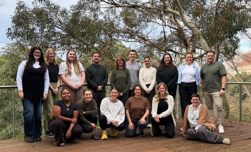 The Zebras team (17 people) posing together with a backdrop of gum trees. People in the front row are crouching, while people in the back are standing up.